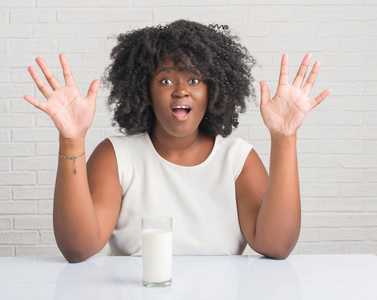 年轻的非裔美国妇女坐在桌子上喝着一杯牛奶，非常高兴和兴奋，胜利者的表情庆祝胜利，微笑着尖叫，举起双手