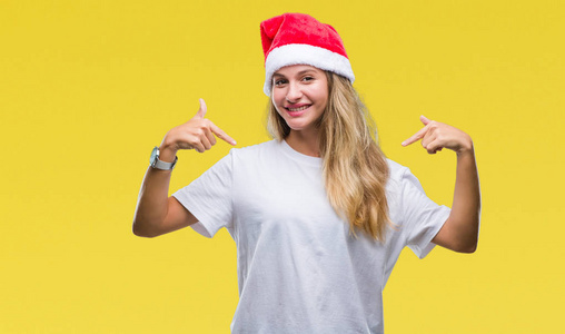 年轻漂亮的金发女人戴着圣诞帽，戴着孤立的背景，脸上带着，用手指指着自己，骄傲和快乐。