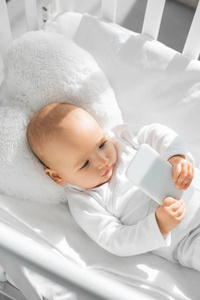 可爱的婴儿婴儿抱着智能手机，白色婴儿床