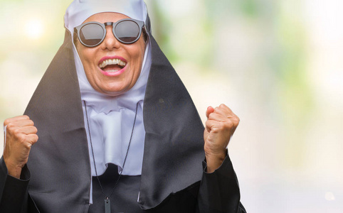 中年资深天主教修女女士戴着太阳镜在孤立的背景上庆祝惊讶和惊讶的成功，举起手臂和睁开眼睛。 赢家概念。