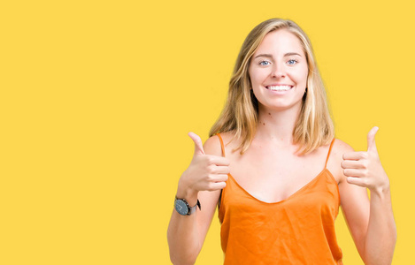 美丽的年轻女子穿着橙色衬衫，在孤立的背景上，成功标志，用手竖起大拇指，微笑和快乐地做积极的手势。 用欢快的表情看着相机，胜利者的