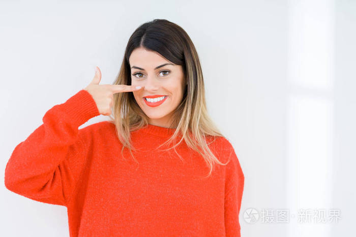年轻女子穿着休闲的红色毛衣背景偏僻用手指着脸和鼻子微笑开朗