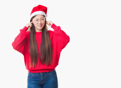 年轻的中国女人在孤立的背景上戴着圣诞帽，用手指遮住耳朵，对响亮的音乐的声音表达出恼怒的表情。 聋的概念。
