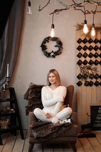 年轻美丽的金发碧眼的女孩在一个温暖的白色针织毛衣的背景下, 一个家庭舒适的房间与圣诞花环