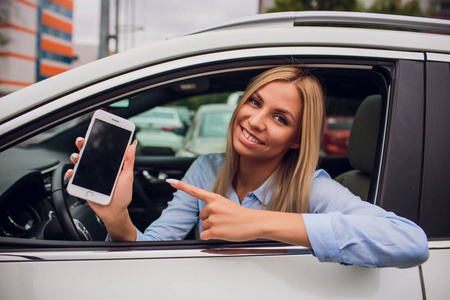 快乐的金发碧眼的女人展示智能手机窗外的汽车。专注于手机