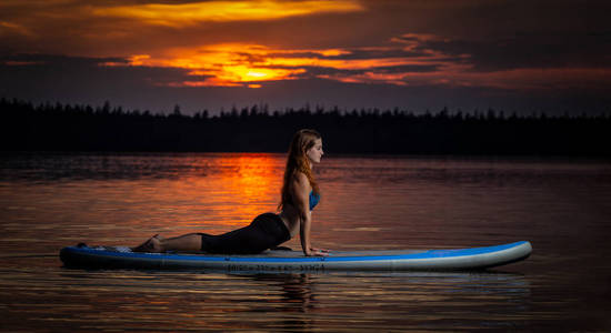 美丽的身材苗条的女孩，长着棕色头发，在黑暗的彩色夕阳下，在风景优美的湖面上，天鹅绒般的黑道上，在桨板上锻炼瑜伽