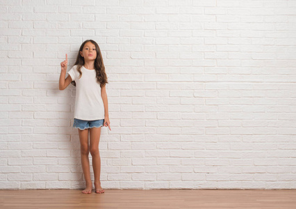 年轻的西班牙裔孩子站在白色砖墙上，用第一个手指指着，同时微笑着自信和快乐。