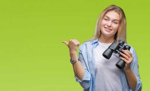 年轻的白种人女人拿着双筒望远镜在孤立的背景上指指点点，用拇指向旁边看，脸上带着幸福的微笑