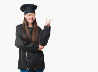 年轻的中国女人在孤立的背景下穿着厨师制服，微笑着，快乐的脸对着镜头眨着眼睛，做着胜利的标志。 二号。