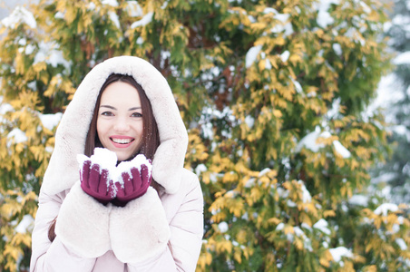 年轻美丽的情感女人的肖像，穿着带帽的羽绒服和手套，在白雪覆盖的花园背景上。 冬季雪景