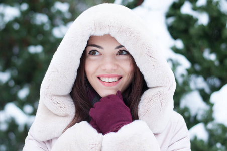 年轻美丽的情感女人的肖像，穿着带帽的羽绒服和手套，在白雪覆盖的花园背景上。 冬季雪景