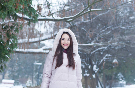 年轻美丽的情感女人的肖像，穿着带帽的羽绒服和手套，在白雪覆盖的花园背景上。 冬季雪景