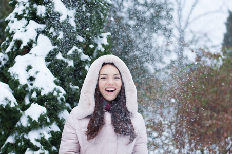 年轻美丽的情感女人的肖像，穿着带帽的羽绒服和手套，在白雪覆盖的花园背景上。 冬季雪景