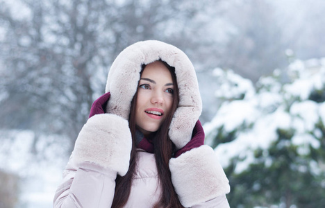 年轻美丽的情感女人的肖像，穿着带帽的羽绒服和手套，在白雪覆盖的花园背景上。 冬季雪景