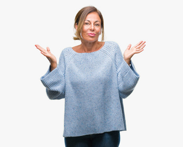 中年老年西班牙裔妇女穿着冬季毛衣，在孤立的背景下毫无头绪，表情混乱，手臂和手举起来。 怀疑概念。