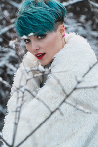 一个蓝色头发的好女孩把自己包裹在一个白雪覆盖的公园里的白色毛皮外套里。 摩擦灌木丛的背景