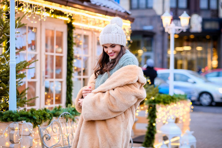 一个穿着冬帽和皮大衣的美丽女孩站在圣诞期间装饰着美丽灯光的晚间街道上