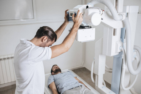 年轻的医疗技术人员操作X光机，而他的病人躺在他面前的床上在一家诊所。