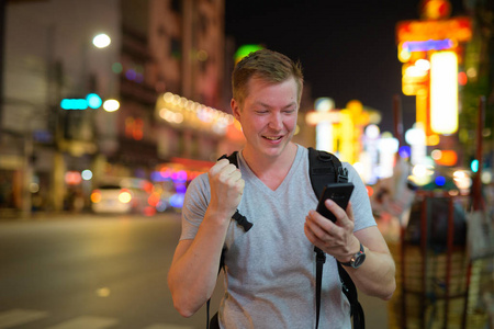 快乐的旅游男子背包客使用电话和得到好消息在唐人街在晚上