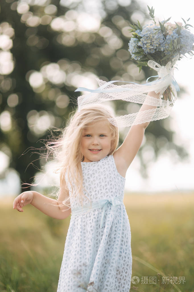 小女孩在天空蓝色礼服与花束站立在领域在大树前面.孩子的微笑和乐趣