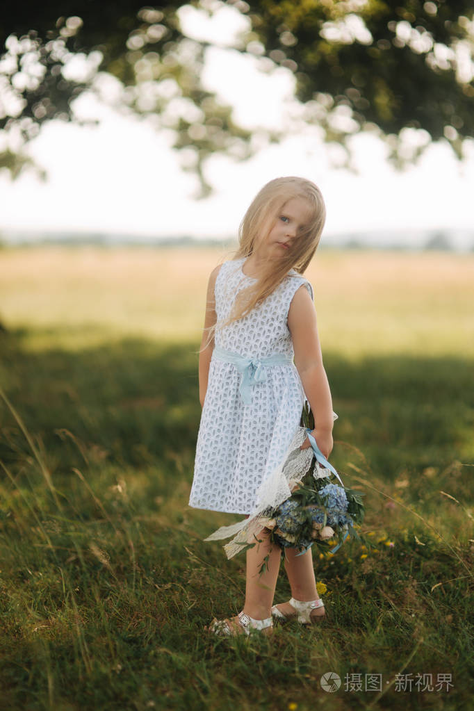 小女孩在天空蓝色礼服与花束站立在领域在大树前面孩子的微笑和乐趣