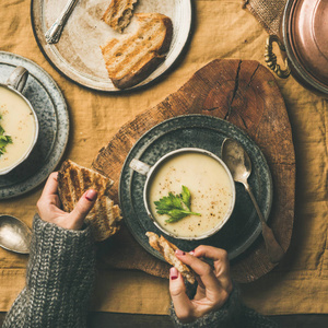 秋天的冬天回家晚餐。 平躺的秋季温暖芹菜奶油汤和女性手与面包上的亚麻桌布，俯视广场作物。 舒适食物素食健康慢食概念