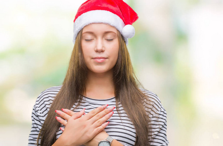 年轻美丽的高加索女人戴着圣诞帽，在孤立的背景上微笑，双手放在胸前，闭着眼睛，脸上有感激的手势。 健康概念。
