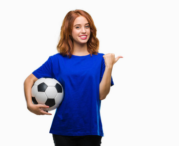 年轻的美女，在孤立的背景下，手里拿着足球球，拇指指向一边，脸上洋溢着幸福的笑容
