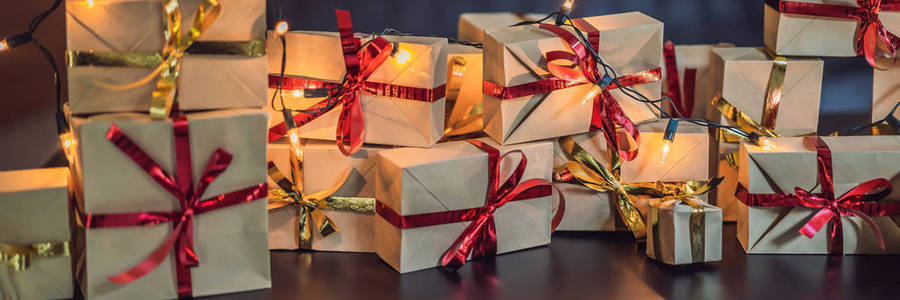一堆礼物包裹在工艺纸上，上面有红色和金色的丝带，放在黑色的木桌上，上面有仙女的灯光，供圣诞假期使用。