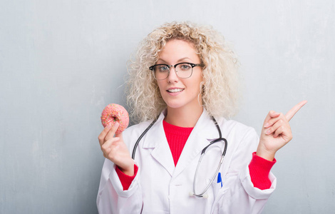年轻的金发碧眼的医生女医生在灰色的墙壁上抱着甜甜圈，非常高兴地用手和手指指向一边