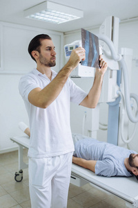 年轻的医疗技术人员检查x射线电影，而他的病人躺在他旁边的床上在一家诊所。