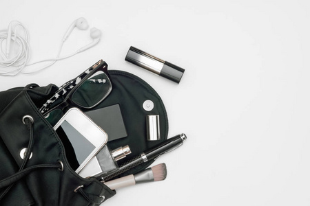 女式黑包的顶部视图，配有配件智能手机香水笔化妆品耳机和白色背景眼镜。