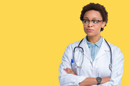 年轻的非裔美国医生女人穿着医用外套，在孤立的背景上微笑着看着一边，凝视着远方的思考。