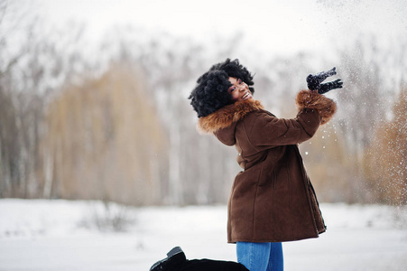 卷发非洲裔美国妇女穿着羊皮外套和手套在冬天的时候摆好姿势，卷起雪。