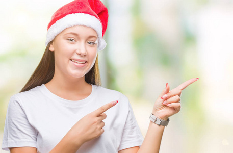 年轻美丽的高加索女人戴着圣诞帽，在孤立的背景上微笑着，看着镜头，双手和手指指向一边。