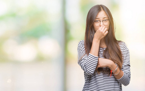 年轻的亚洲女人戴着眼镜，在孤立的背景上闻着一些臭味和令人恶心的难以忍受的气味，用手指在鼻子上屏住呼吸。 难闻的概念。