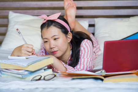 年轻，可爱，快乐，书呆子，亚洲学生，十几岁的女孩，穿着发带，在家里学习，卧室里坐在床上，拿着笔记本电脑和教科书，微笑着，自信地微