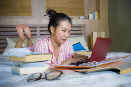年轻漂亮聪明的亚裔美国学生女孩在家卧室里坐在床上，用笔记本电脑和，微笑着自信地看待大学教育概念