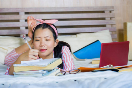 年轻，可爱，快乐，书呆子，亚洲学生，十几岁的女孩，穿着发带，在家里学习，卧室里，坐在床上，笔记本电脑和教科书，微笑，自信，自信