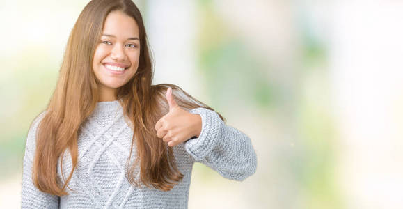 年轻漂亮的黑发女人穿着毛衣，在孤立的背景下，用手做快乐的竖起大拇指的手势。 赞许的表情看着相机，显示出成功。
