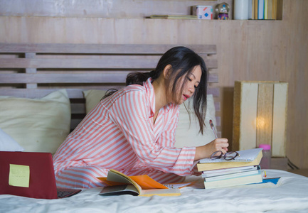 年轻漂亮聪明的亚洲学生女孩在家卧室里坐在床上，用笔记本电脑和教科书进行考试，专注于教育概念