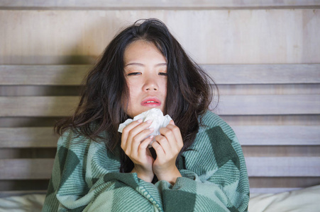年轻美丽甜美的亚洲女孩，穿着睡衣，裹着毯子，患感冒和流感，在床上体温计，感到发烧，吹着鼻子咳嗽