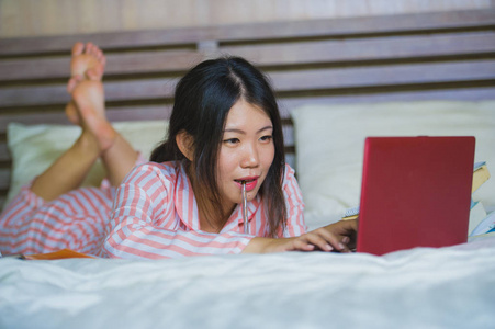 年轻漂亮聪明的亚裔韩国学生女孩在家卧室里坐在床上，用笔记本电脑和教科书进行考试，微笑着对大学和教育的概念充满信心