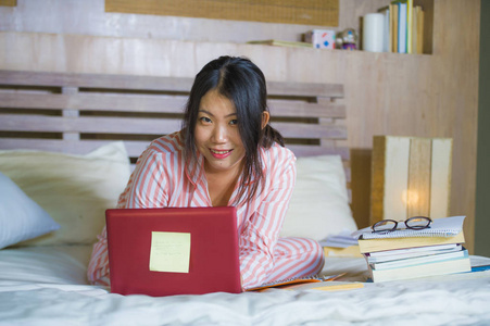 年轻漂亮聪明的亚裔韩国女学生在家，卧室，坐在床上，用笔记本电脑和教科书进行考试，微笑着对大学和教育概念充满信心