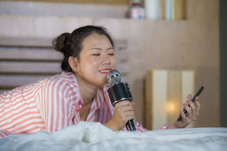 年轻快乐美丽的亚裔美国少年女孩唱卡拉OK歌兴奋在家卧室拿着手机在床上玩兴奋和欢快的模仿流行音乐明星