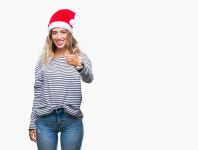 美丽的年轻金发女人戴着圣诞帽，在孤立的背景上，用手做快乐的竖起大拇指的手势。 赞许的表情看着相机，显示出成功。