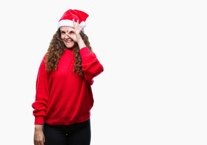 年轻的黑发女孩戴着圣诞帽在孤立的背景上做着好的手势，用手微笑的眼睛透过手指看着快乐的脸。