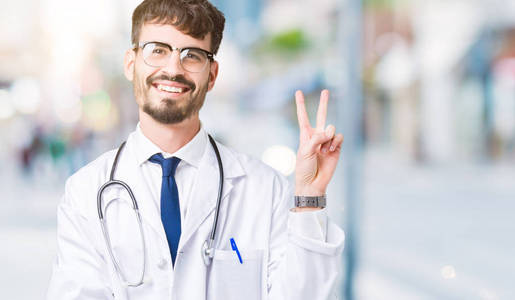 年轻的医生穿着医院外套，在孤立的背景上微笑，快乐的脸对着镜头眨眼，做胜利的标志。 二号。