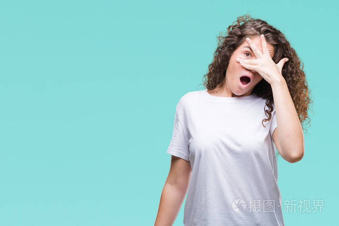 美丽的黑发卷发年轻女孩穿着休闲t恤,在孤立的背景上偷看震惊,覆盖着
