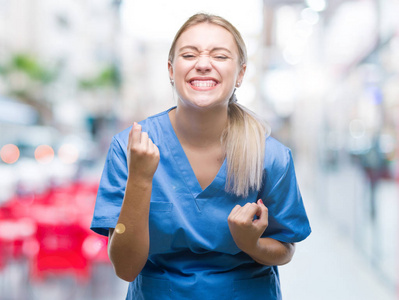 年轻的金发外科医生女士穿着医疗制服，在孤立的背景下，非常高兴和兴奋地做着胜利者的手势，举起手臂，微笑着，尖叫着，争取成功。 庆祝